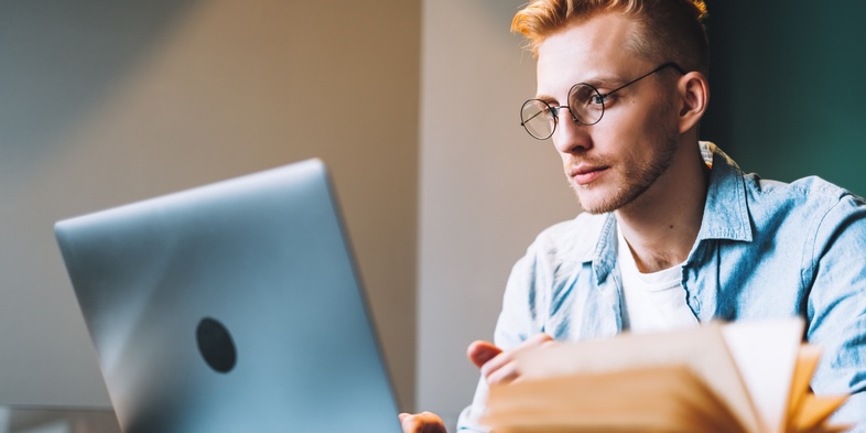 Ung man med glasögon studerar vid en bärbar dator och har en uppslagen bok framför sig.