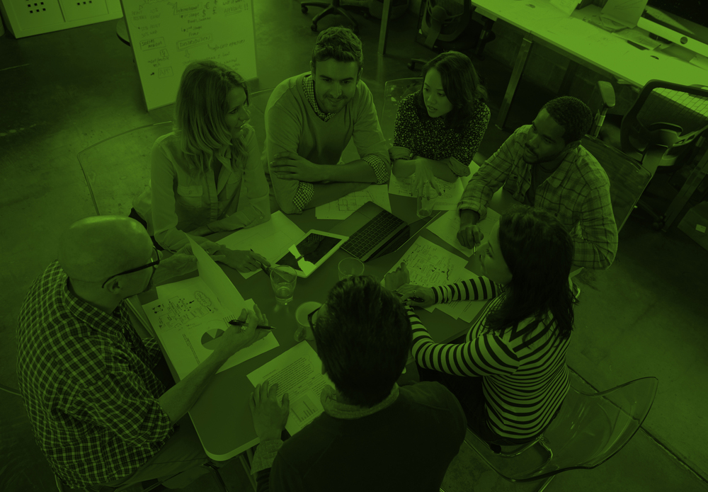 En grupp människor sitter runt ett bord och har ett möte på ett kontor, bilden har ett grönt filter.