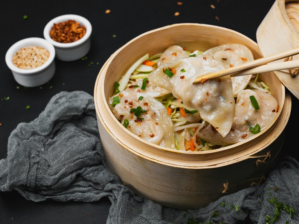 En bambukorg fylld med ångade dumplings ovanpå strimlade grönsaker, med ätpinnar och små skålar med kryddor bredvid.
