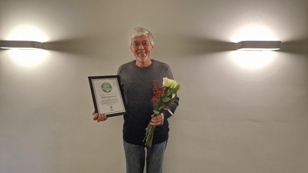 En äldre man står mot en ljus vägg, håller en bukett blommor och ett diplom, och ler mot kameran.