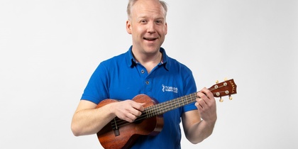 Man i blå pikétröja spelar ukulele och står mot en vit bakgrund.