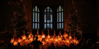 En grupp människor i vita kläder håller ljus i en mörk kyrka, omgivna av julgranar med ljusslingor.