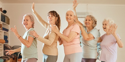 Fem äldre kvinnor gör yoga eller dansar tillsammans i ett ljust träningsrum med trägolv.
