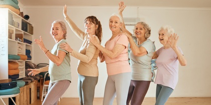 Fem äldre kvinnor gör yoga eller dansar tillsammans i ett ljust träningsrum med trägolv.
