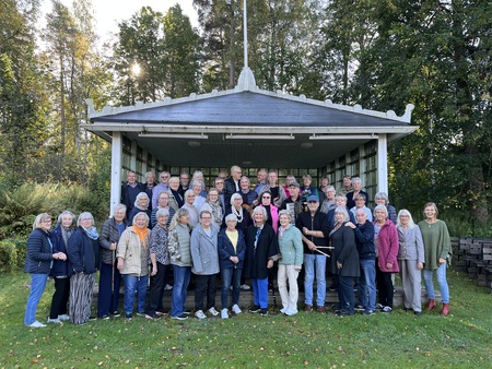 En stor grupp människor står utomhus framför en liten scen i en grönskande park.