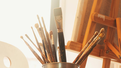 En burk med olika penslar står framför ett staffli i ett ljust konstnärsrum.