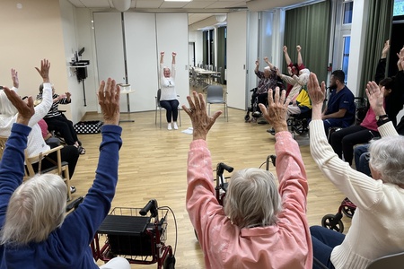 En grupp äldre personer sitter i en cirkel och gör sittande gymnastik med armarna uppsträckta.