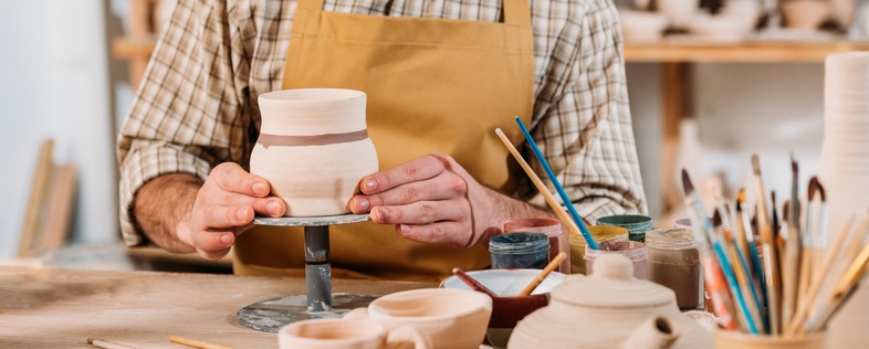 En person i förkläde arbetar med keramik på en drejskiva i en verkstad fylld med lergods och penslar.