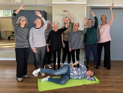 En grupp äldre kvinnor poserar glatt i ett träningsrum, en kvinna ligger på en grön yogamatta framför de andra.