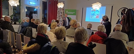 En grupp äldre personer sitter i ett rum och lyssnar på en presentation med bildspel.