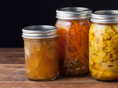 Tre glasburkar med inlagda grönsaker och frukt står på ett träbord mot en mörk bakgrund.