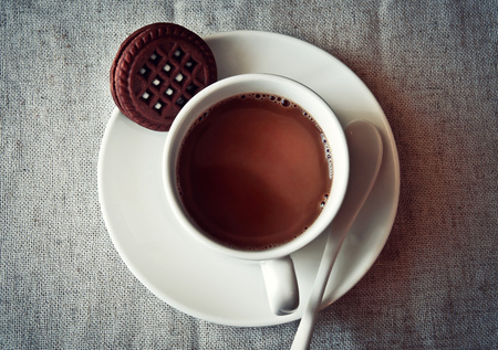 En kopp varm choklad på ett vitt fat med en chokladkaka och en vit sked, ovanifrån.