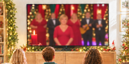 En grupp människor sitter i ett vardagsrum och tittar på ett julprogram på TV, omgivna av juldekorationer.