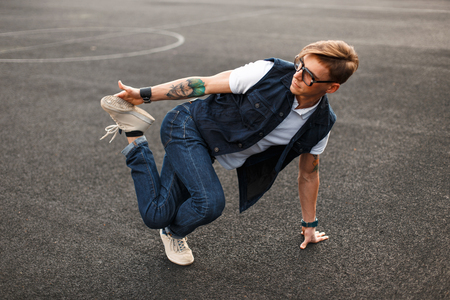 En person dansar breakdance på asfalt, klädd i jeans, vit t-shirt och glasögon.
