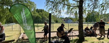 En person spelar gitarr och sjunger utomhus under ett träd, med åhörare i bakgrunden och en grön flagga vid sidan.