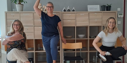 Tre kvinnor poserar lekfullt på och bredvid stolar i ett ljust kontorsrum med hyllor i bakgrunden.