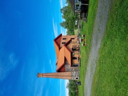 Gammal tegelbyggnad med skorsten på landsbygden under klarblå himmel, omgiven av gräs och träd.