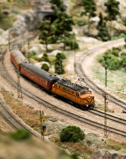 Modelltåg i orange färg kör på en kurvig järnväg genom ett landskap med träd och grönska.