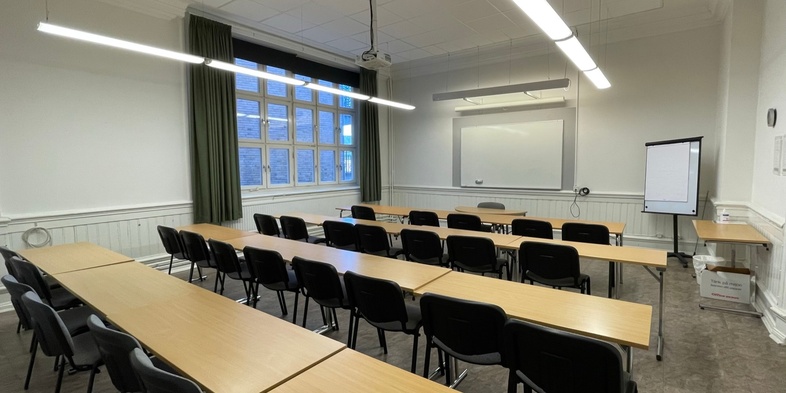 Ett tomt klassrum med rader av bord och stolar, en whiteboard, fönster och en blädderblockstavla.