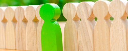 En grön träfigur står mellan två rader av ljusa träfigurer på ett bord med grön bakgrund.