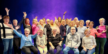 En stor grupp glada vuxna poserar tillsammans framför en lila och blå bakgrund, flera med armarna i luften.
