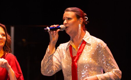 En person i glittrig skjorta och röd slips sjunger i mikrofon på scen.