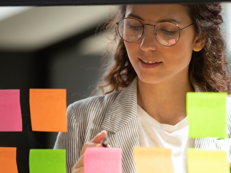 Kvinna med glasögon skriver på färgglada post-it-lappar på en glasvägg på ett kontor.