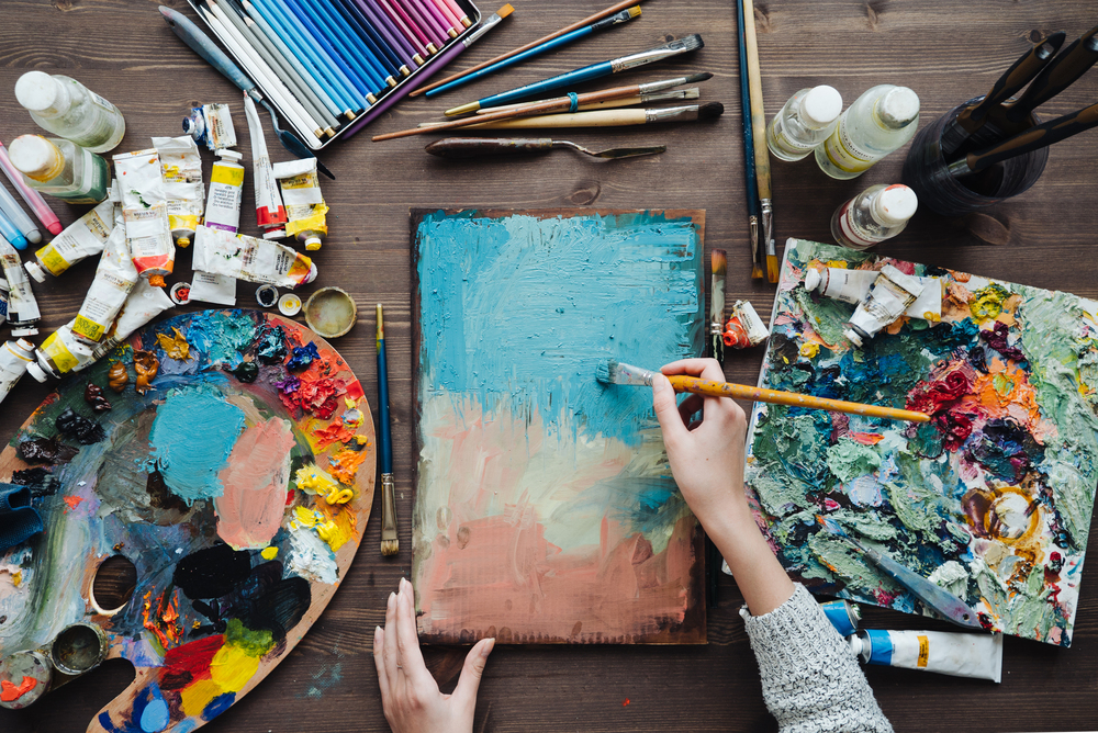 En person målar abstrakt konst på en duk med pensel, omgiven av färgtuber, penslar och färgpaletter på ett bord.