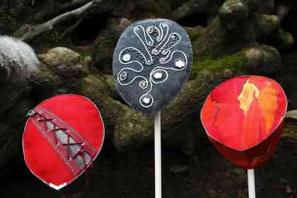 Tre färgglada, handgjorda masker på pinnar mot en bakgrund av mossa och stenar.