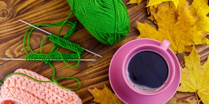 Stickning med grönt garn, rosa stickat plagg, rosa kaffekopp och gula höstlöv på ett träbord.