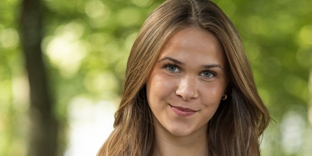 Ung kvinna med långt brunt hår och vit t-shirt står utomhus i en grönskande park.