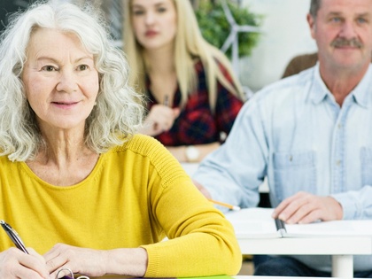 Äldre kvinna med grått hår sitter i klassrum och skriver, andra personer syns i bakgrunden.
