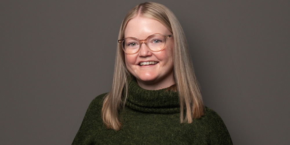 Person med långt blont hår, grön stickad tröja och svarta byxor står mot en grå bakgrund.