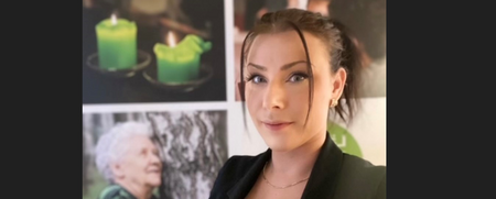 Kvinna i svart kavaj står framför en affisch med gröna ljus, äldre kvinna och växter.