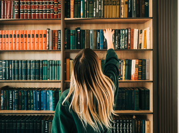 En person med långt blont hår sträcker sig efter en bok på en bokhylla fylld med böcker.