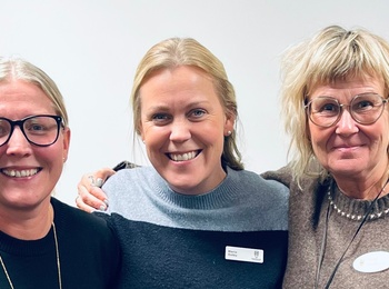 Tre personer står tillsammans i ett ljust rum, klädda i stickade tröjor och med namnskyltar.
