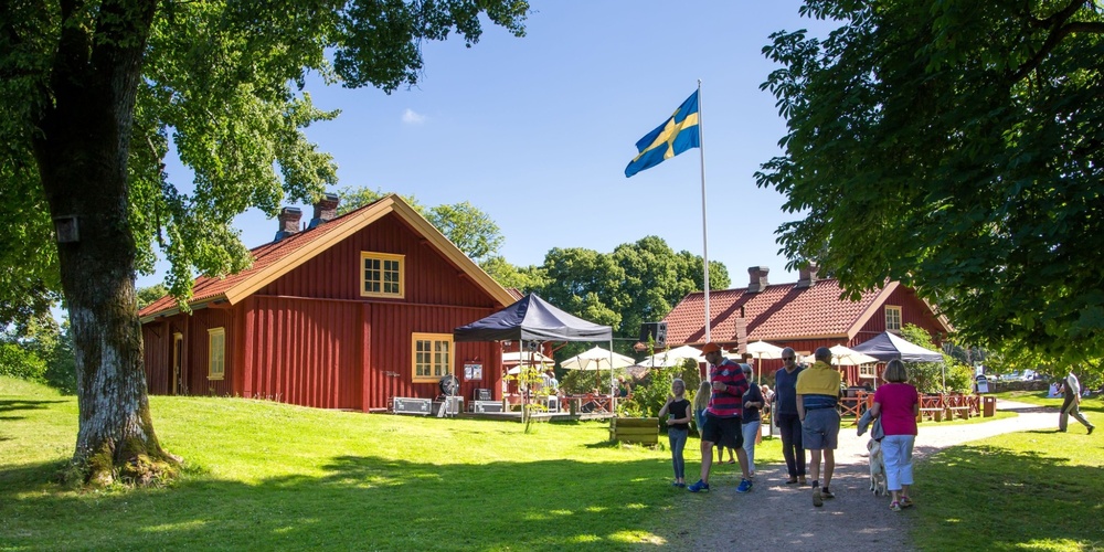 Folk promenerar mot röda trähus med svensk flagga, soligt väder och grönskande omgivning.