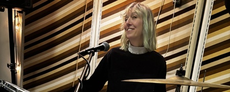 En leende person sitter vid ett trumset i en musikstudio med randig vägg bakom.