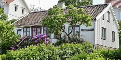 Vit trävilla med blommande buskar och trädgård, sommargrönska och grusgång framför huset.