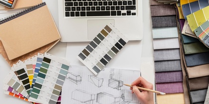 Person skissar möbler vid ett skrivbord med tygprover, färgkartor och en laptop.