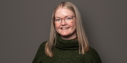 Person med långt blont hår, grön stickad tröja och svarta byxor står mot en grå bakgrund.