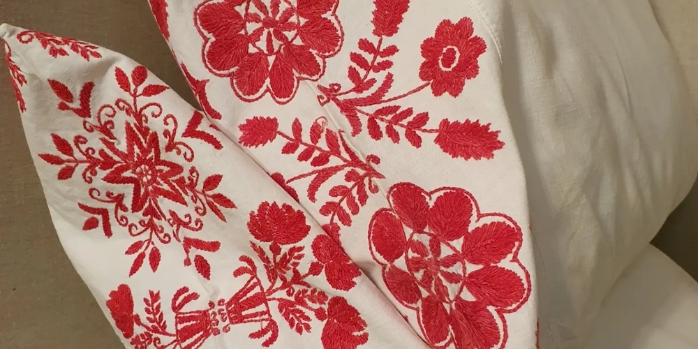 Två vita kuddar med rött, blommigt broderimönster ligger mot en beige bakgrund.