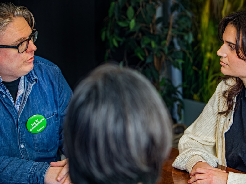 Tre personer sitter vid ett bord och samtalar, två ansikten synliga, en person med ryggen mot kameran.