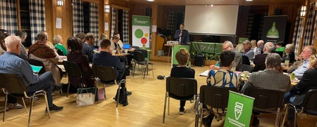 En grupp människor sitter i ett träpanelat rum och lyssnar på en talare vid ett podium.