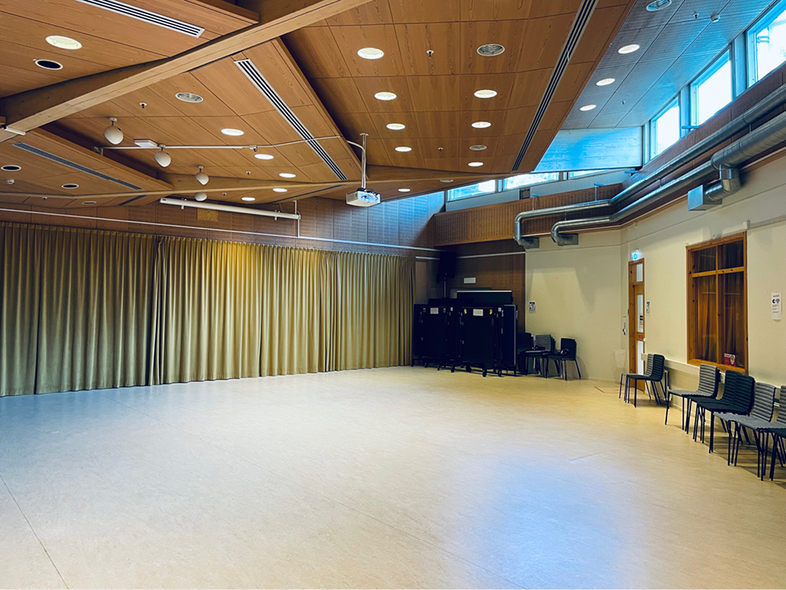 Ljus och tom sal med trägolv, högt i tak, stora fönster och stolar staplade längs väggen.