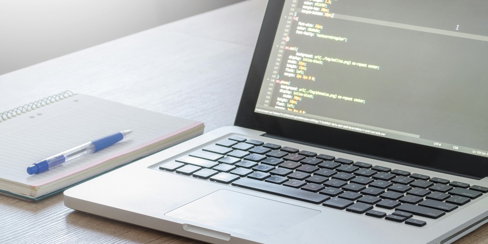 En bärbar dator med kod på skärmen, ett anteckningsblock och en penna på ett skrivbord.