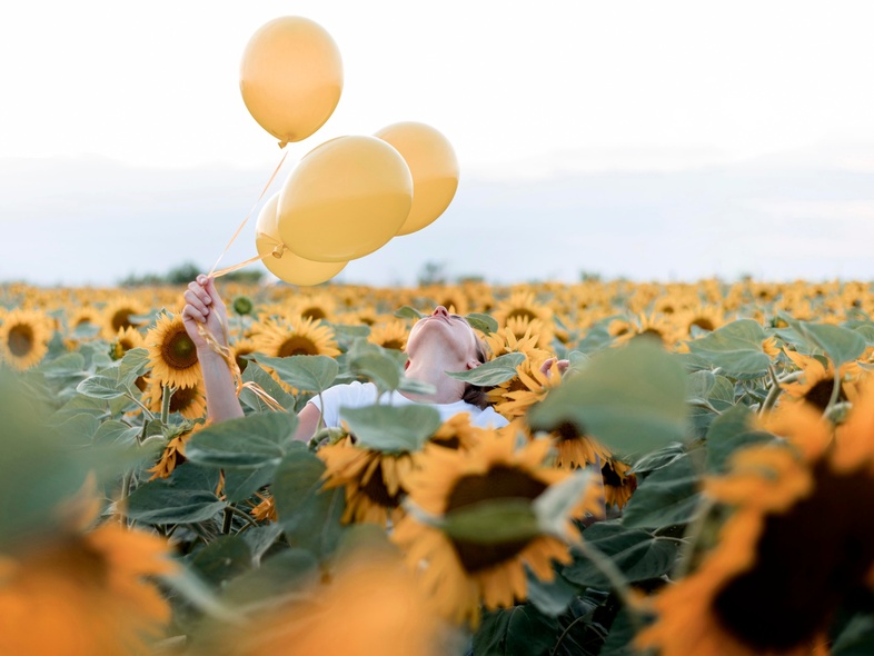 En person håller tre gula ballonger i ett fält av solrosor under en ljus himmel.
