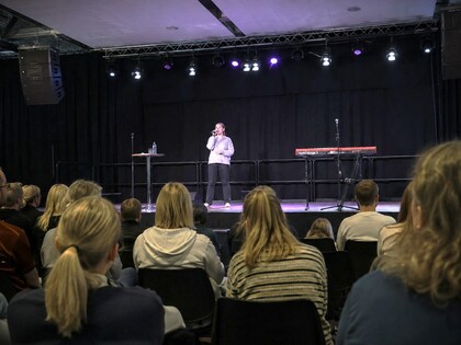 En person står på en scen och talar inför en sittande publik i en mörk sal med scenbelysning.