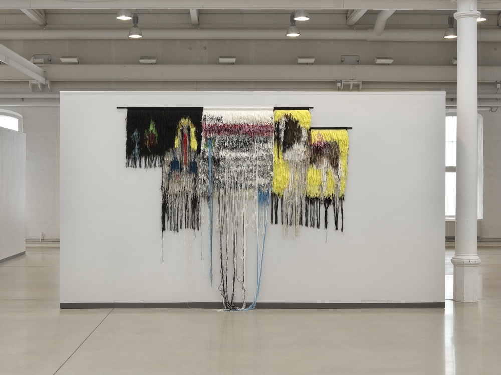 Textil konstinstallation med färgglada trådar som hänger från en vägg i ett galleri.