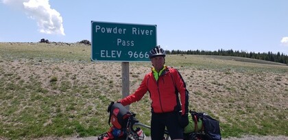 Cyklist i röd jacka står vid en skylt för Powder River Pass, höjd 9666 fot, med cykel och packning.